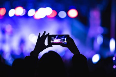 Elinde akıllı telefon ve sahne ışıkları olan konser kalabalığının siluetlerini tutan kişi