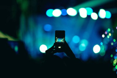 Elinde akıllı telefon ve sahne ışıkları olan konser kalabalığının siluetlerini tutan kişi