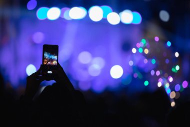 Elinde akıllı telefon ve sahne ışıkları olan konser kalabalığının siluetlerini tutan kişi