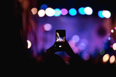 Elinde akıllı telefon ve sahne ışıkları olan konser kalabalığının siluetlerini tutan kişi