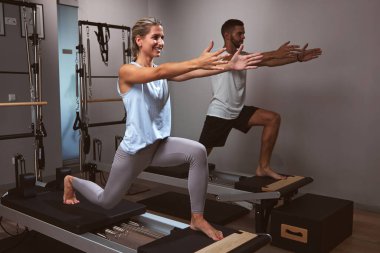 Genç bir kadın ve erkek spor salonunda pilates makinesinde kişisel antrenörleriyle egzersiz yapıyorlar..