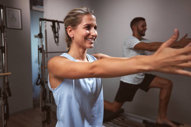 Genç bir kadın ve erkek spor salonunda pilates makinesinde kişisel antrenörleriyle egzersiz yapıyorlar..