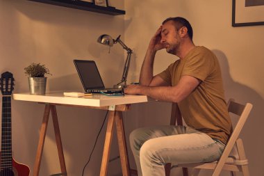 Evde dizüstü bilgisayarla uğraşırken başı ağrıyan ve bitkin hisseden bir adam..