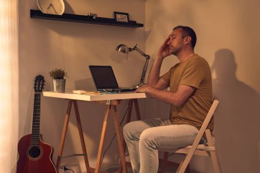 Evde dizüstü bilgisayarla uğraşırken başı ağrıyan ve bitkin hisseden bir adam..