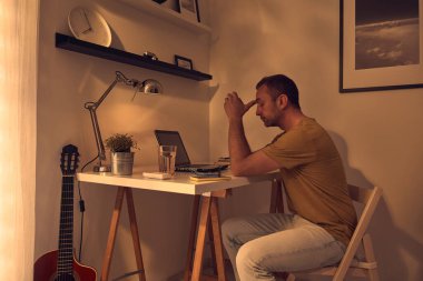 Evde dizüstü bilgisayarla uğraşırken başı ağrıyan ve bitkin hisseden bir adam..