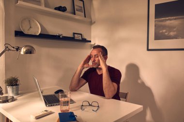 Evde dizüstü bilgisayarla uğraşırken başı ağrıyan ve bitkin hisseden bir adam..