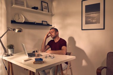 Evde dizüstü bilgisayarla uğraşırken başı ağrıyan ve bitkin hisseden bir adam..