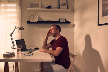 Evde dizüstü bilgisayarla uğraşırken başı ağrıyan ve bitkin hisseden bir adam..