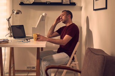 Evde dizüstü bilgisayarla uğraşırken başı ağrıyan ve bitkin hisseden bir adam..