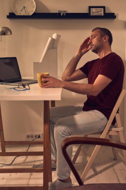 Evde dizüstü bilgisayarla uğraşırken başı ağrıyan ve bitkin hisseden bir adam..