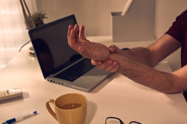 Evde dizüstü bilgisayarla çalışırken eli ve kolu ağrıyan bir adam..