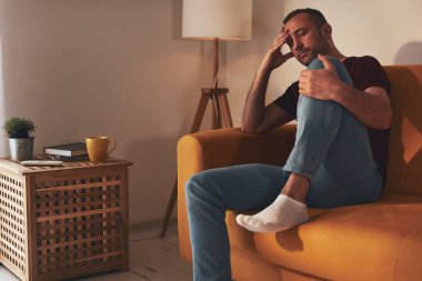 Baş ağrısı ve diz ağrısı olan bir adam evdeki kanepede oturuyor..