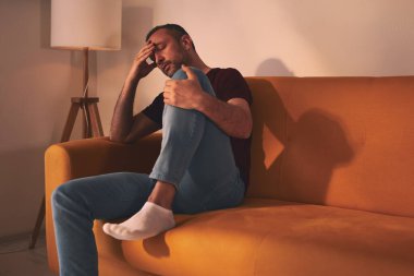 Baş ağrısı ve diz ağrısı olan bir adam evdeki kanepede oturuyor..
