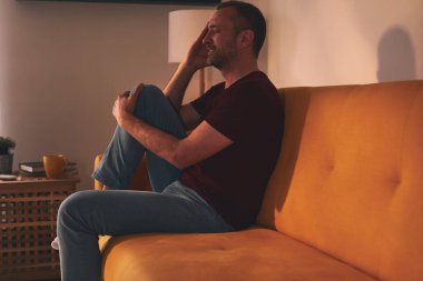 Baş ağrısı ve diz ağrısı olan bir adam evdeki kanepede oturuyor..