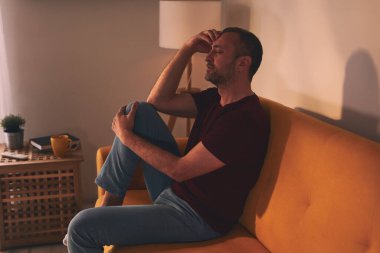 Baş ağrısı ve diz ağrısı olan bir adam evdeki kanepede oturuyor..