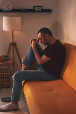 Baş ağrısı ve diz ağrısı olan bir adam evdeki kanepede oturuyor..
