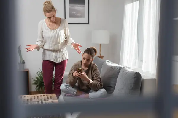 Mother having trouble with a lazy teenager girl at home while playing video games on a smartphone.