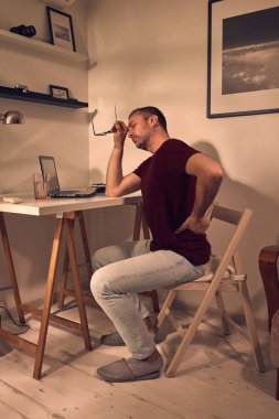 Evde dizüstü bilgisayar üzerinde çalışırken baş ve sırt ağrısı çeken bir adam..