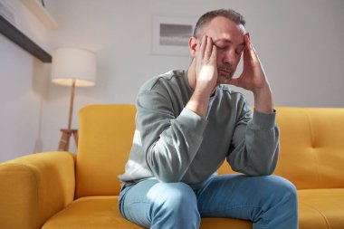 Başı ağrıyan adam evdeki kanepede oturuyor..