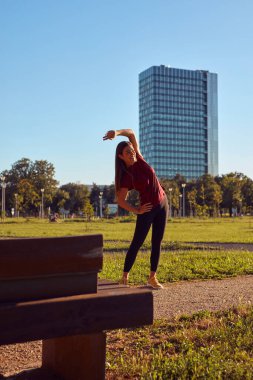 Genç bir kadın güneşli yaz gününde şehir parkında egzersiz ve koşu için ısınıyor..