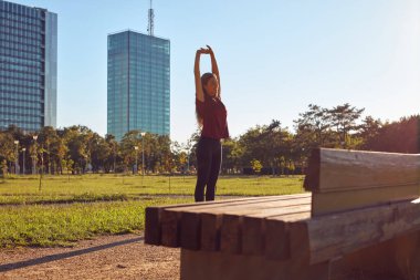 Genç bir kadın güneşli yaz gününde şehir parkında egzersiz ve koşu için ısınıyor..