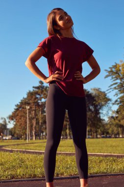 Genç bir kadın güneşli yaz gününde şehir parkında egzersiz ve koşu için ısınıyor..