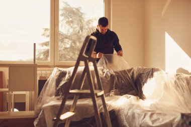 Evdeki kanepeyi yenileme sırasında tozdan korunmak için plastik çarşaflarla örten adam..