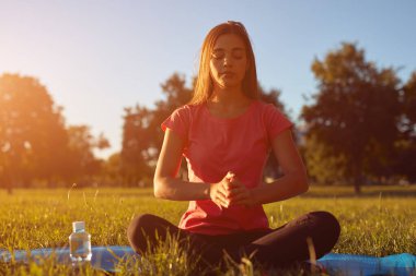 Young woman exercising on a sports yoga mat on the grass in a park during summertime sunset time. clipart