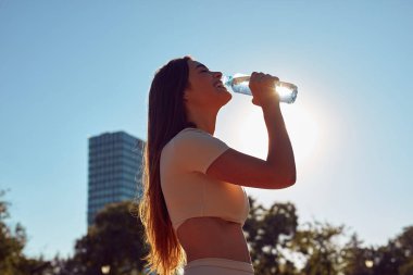 Şehir parkında koşudan ve egzersizden sonra su içen genç bir kadın.