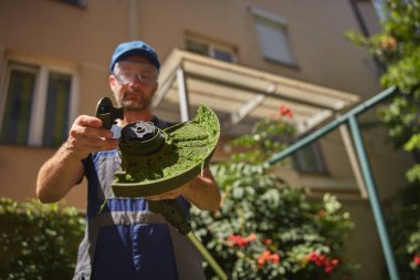 Bahçıvan elinde çim biçme makinesiyle binanın etrafındaki çimleri ve otları biçiyor ve yıpranmış ipleri değiştiriyor..