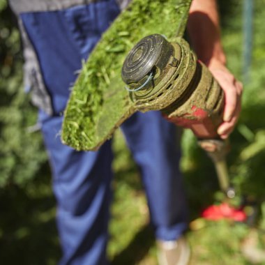 Bahçıvan elinde çim biçme makinesiyle binanın etrafındaki çimleri ve otları biçiyor ve yıpranmış ipleri değiştiriyor..