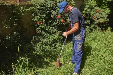 Bahçıvan elinde çim biçme makinesiyle Binanın etrafındaki otları ve otları biçiyor.