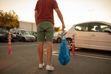Alışveriş çantalı bir adam gün batımında bir marketin otoparkında.