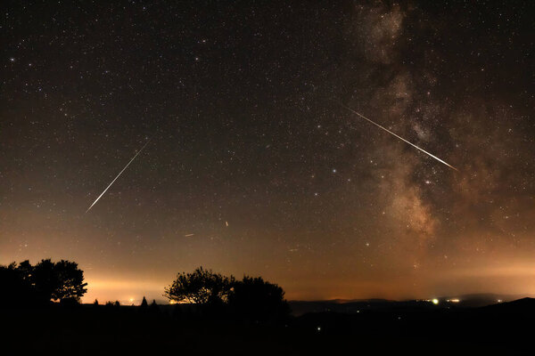 Milky Way stars with meteor shower trails and countryside silhouettes.