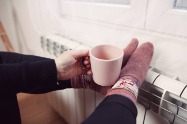 Soğuk bir kış gününde, sıcak çay içen bir kadın, çoraplarıyla evde ısıtma radyatöründe çoraplarını giyiyor..