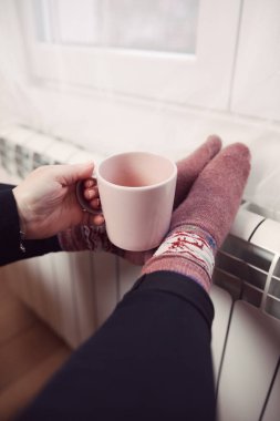 Soğuk bir kış gününde, sıcak çay içen bir kadın, çoraplarıyla evde ısıtma radyatöründe çoraplarını giyiyor..