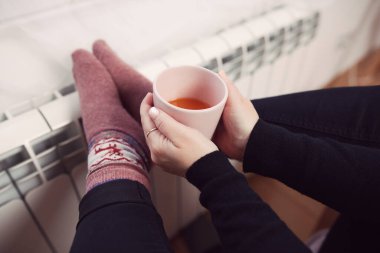 Soğuk bir kış gününde, sıcak çay içen bir kadın, çoraplarıyla evde ısıtma radyatöründe çoraplarını giyiyor..