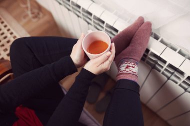 Soğuk bir kış gününde, sıcak çay içen bir kadın, çoraplarıyla evde ısıtma radyatöründe çoraplarını giyiyor..