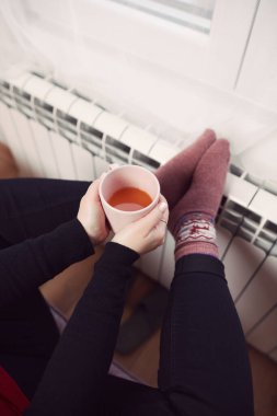 Soğuk bir kış gününde, sıcak çay içen bir kadın, çoraplarıyla evde ısıtma radyatöründe çoraplarını giyiyor..
