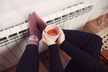 Soğuk bir kış gününde, sıcak çay içen bir kadın, çoraplarıyla evde ısıtma radyatöründe çoraplarını giyiyor..