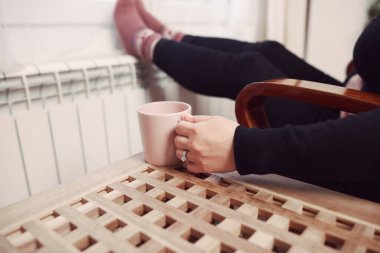 Soğuk bir kış gününde, sıcak çay içen bir kadın, çoraplarıyla evde ısıtma radyatöründe çoraplarını giyiyor..