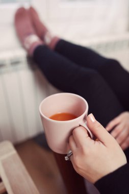 Soğuk bir kış gününde, sıcak çay içen bir kadın, çoraplarıyla evde ısıtma radyatöründe çoraplarını giyiyor..