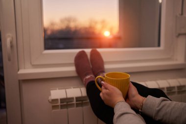 Soğuk bir kış gününde sıcak çay içen bir kadın, çoraplarını giymiş, radyatörünü ısıtıyor..
