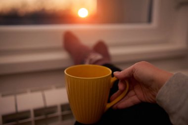 Soğuk bir kış gününde sıcak çay içen bir kadın, çoraplarını giymiş, radyatörünü ısıtıyor..