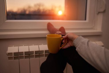 Soğuk bir kış gününde sıcak çay içen bir kadın, çoraplarını giymiş, radyatörünü ısıtıyor..