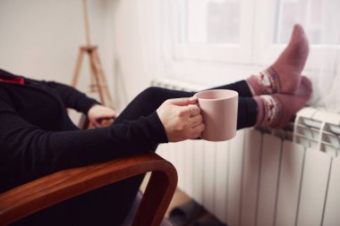 Soğuk bir kış gününde, sıcak çay içen bir kadın, çoraplarıyla evde ısıtma radyatöründe çoraplarını giyiyor..