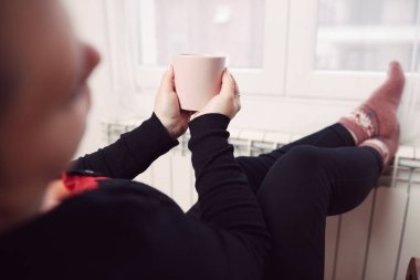 Soğuk bir kış gününde, sıcak çay içen bir kadın, çoraplarıyla evde ısıtma radyatöründe çoraplarını giyiyor..