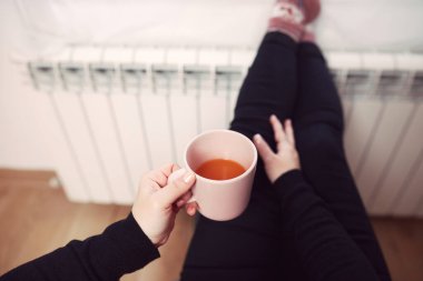 Soğuk bir kış gününde, sıcak çay içen bir kadın, çoraplarıyla evde ısıtma radyatöründe çoraplarını giyiyor..
