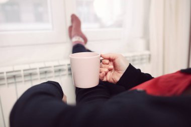 Soğuk bir kış gününde, sıcak çay içen bir kadın, çoraplarıyla evde ısıtma radyatöründe çoraplarını giyiyor..