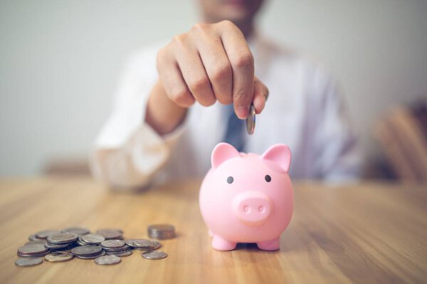 Person in a business shirt saving money in a piggy bank, with coins and financial reports on the table. Saving money business concept.